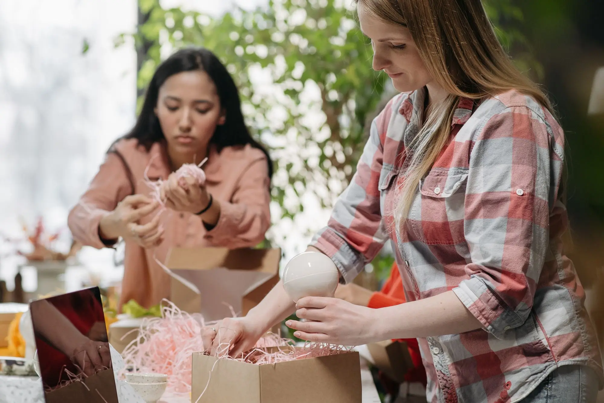 Two founders packing online orders into cardboard boxes with shredded paper filler at their studio table